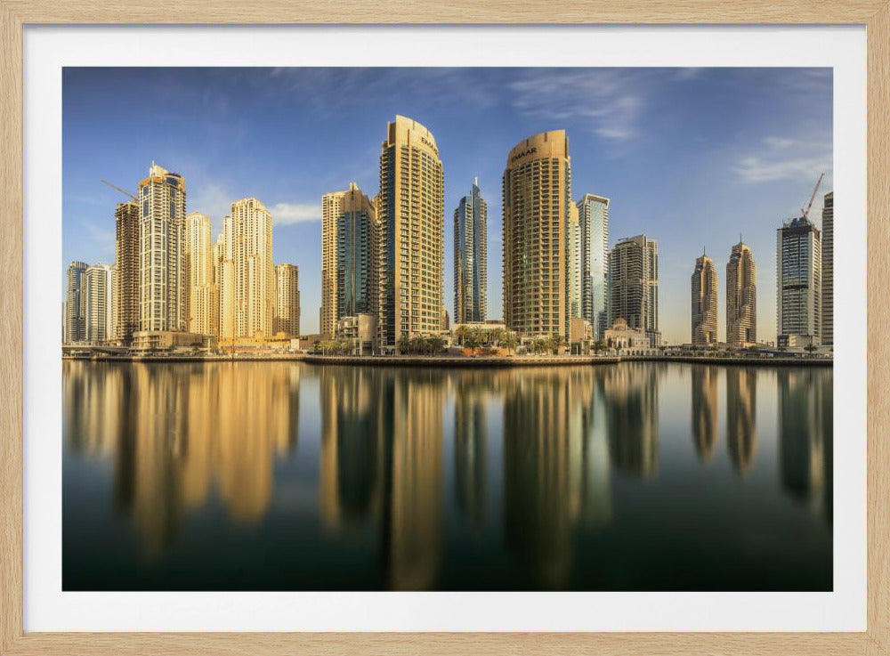 A panoramic photograph of the Dubai Marina skyline with its golden skyscrapers reflecting in the calm, dark water below, all under a clear blue sky and framed in silver. Wall Art