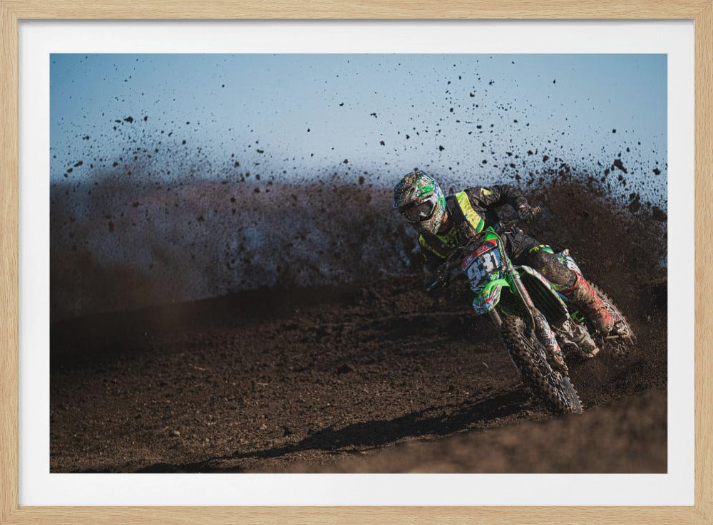 A motocross rider in green and black gear leans hard into a turn on a dirt track, kicking up a large spray of mud against a clear blue sky. Decor