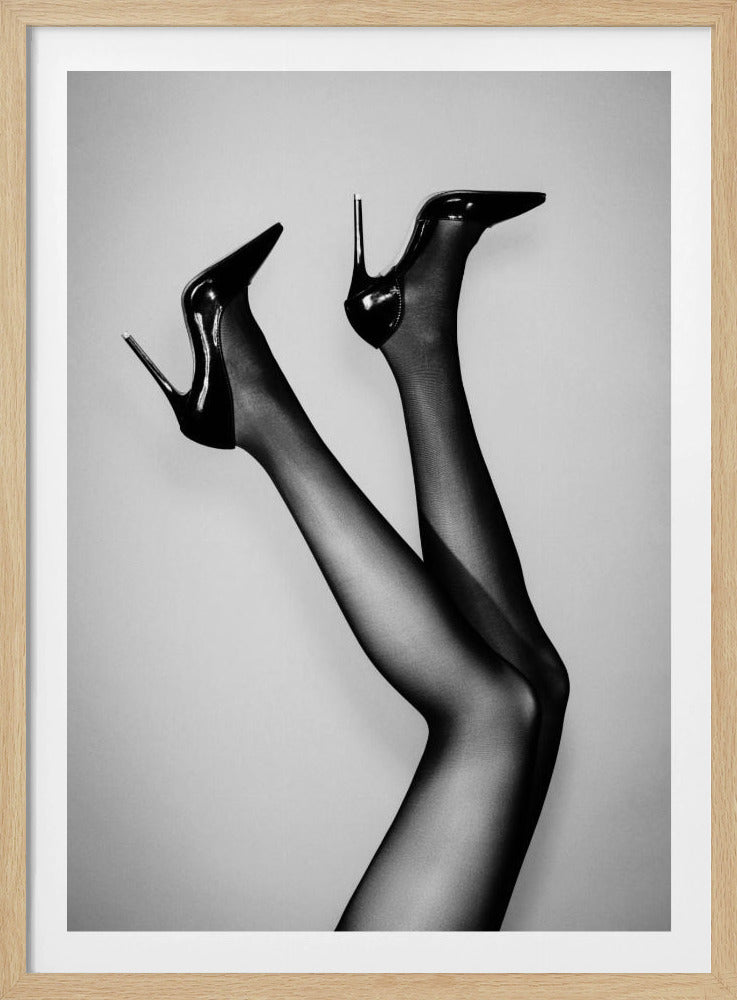 A black and white photograph of a woman's legs in the air, wearing sheer black stockings and shiny black stiletto high heels against a plain grey background. Poster