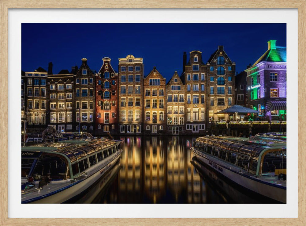 A vibrant night-time photograph of a canal in Amsterdam, flanked by two tour boats in the foreground. Traditional Dutch canal houses are brightly lit, casting warm yellow reflections on the dark water under a deep blue sky. Print