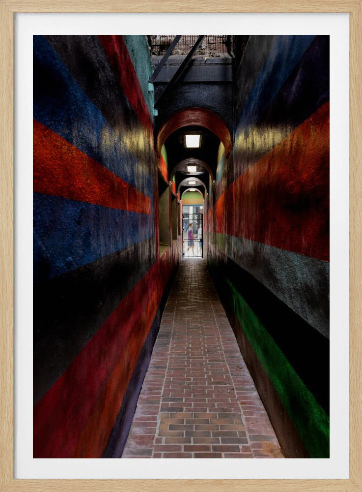A long, narrow brick alleyway viewed from a one-point perspective, with walls painted in wide, colorful horizontal stripes of red, blue, and black, leading to a distant, sunlit exit where a person stands. Wall Art