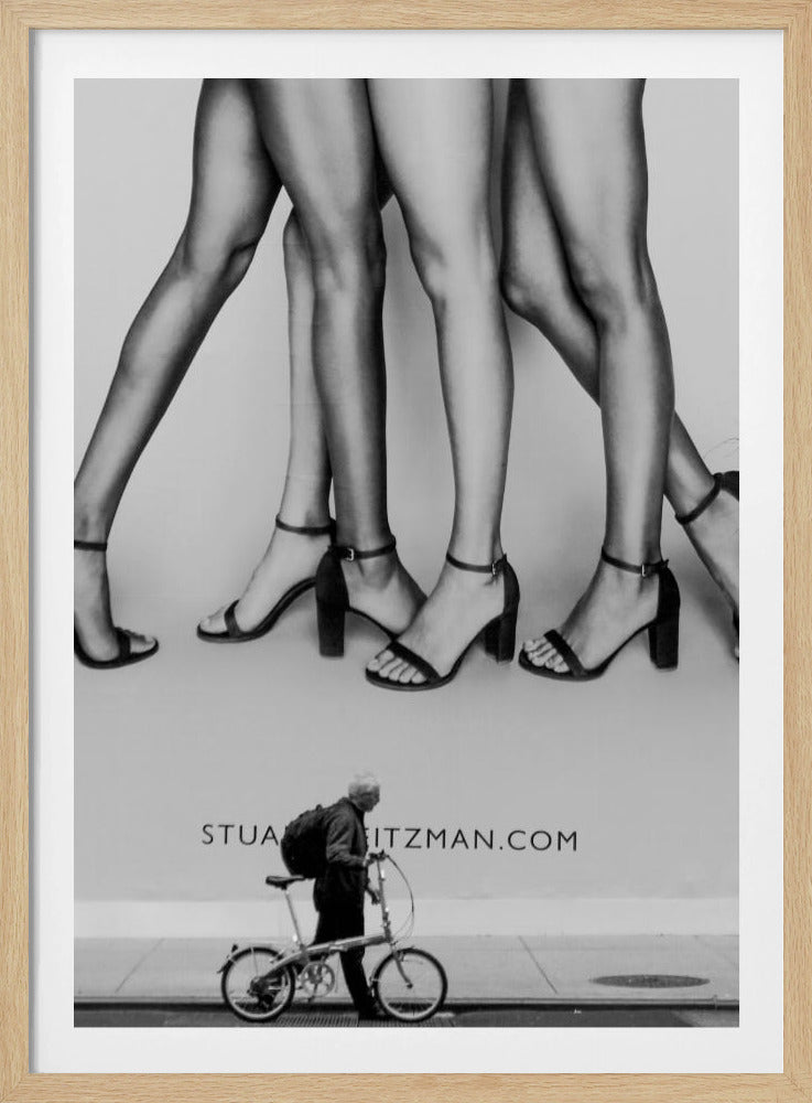A black and white street photograph showing a stark contrast between a large fashion advertisement and a person on the sidewalk. The ad features multiple pairs of long female legs wearing high-heeled sandals. Below the ad, an older man with a backpack walks by, pushing a small foldable bicycle. Artwork