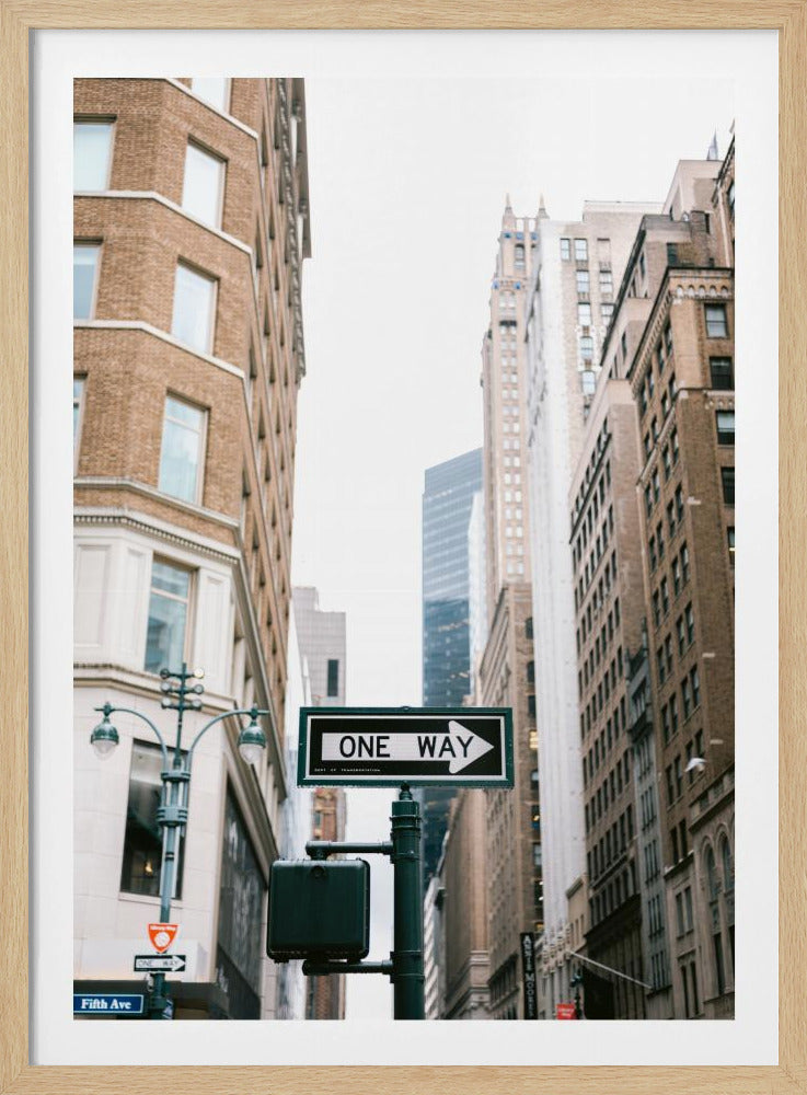 A framed photograph of a New York City street, with a prominent 'ONE WAY' sign in the foreground pointing right. The view is from a low angle, looking up between tall brick and stone buildings towards a bright, overcast sky. Wall Art