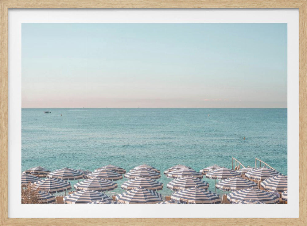 A high-angle photograph of a beach packed with blue and white striped umbrellas, with the turquoise ocean stretching out to a pale blue sky in the background. The entire image is enclosed in a silver frame. Poster