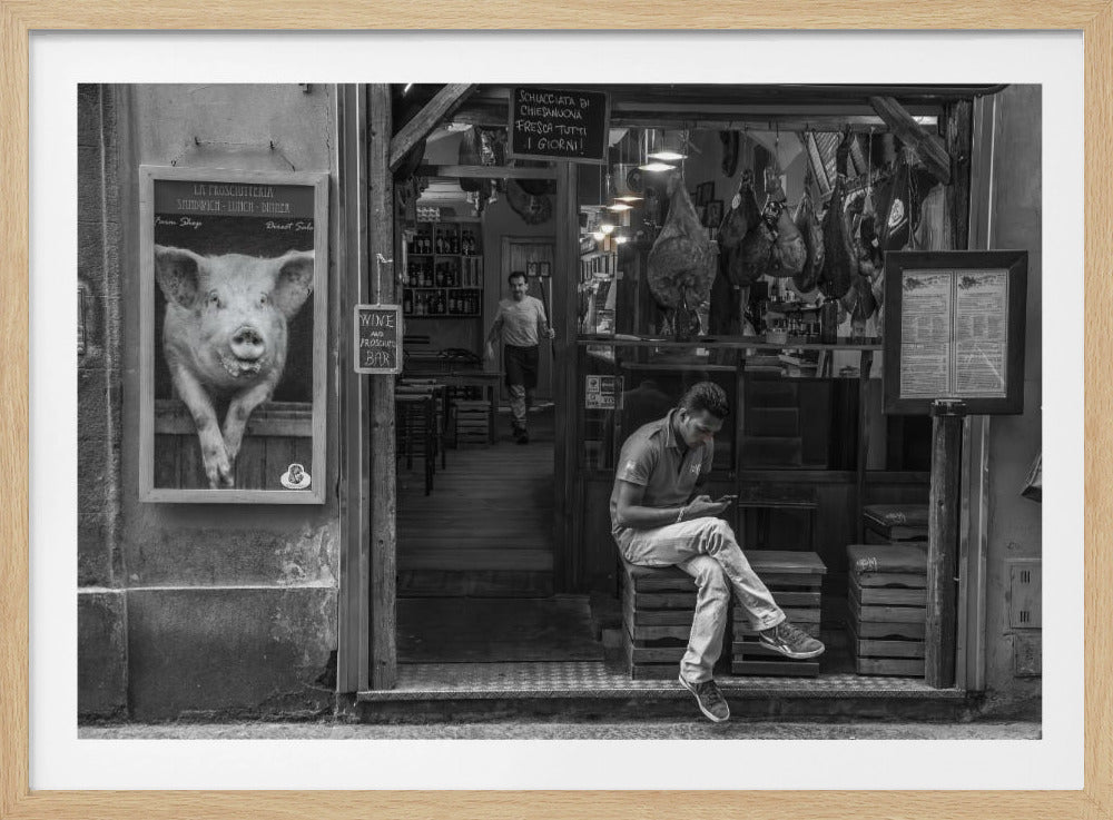 A framed, black and white street photograph of an Italian prosciutto shop. A young man sits on a wooden crate outside, looking at his phone, while cured hams hang inside the shop and a large, framed picture of a pig is on the exterior wall. Artwork