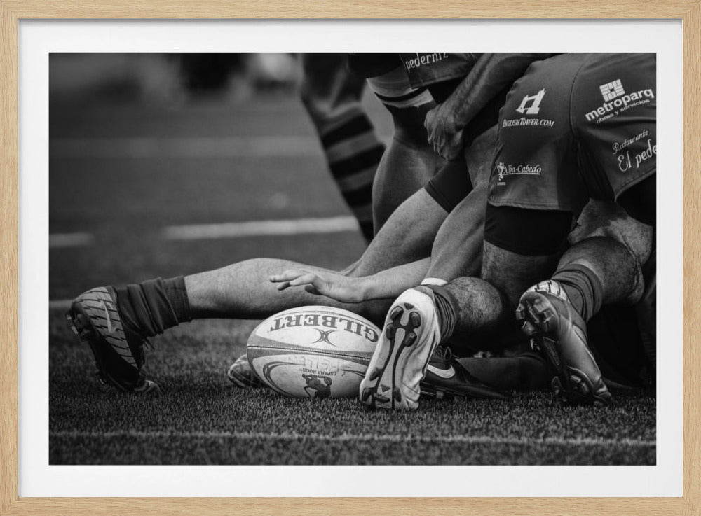 A dynamic black and white, low-angle photograph capturing the intensity of a rugby ruck. A Gilbert rugby ball sits on the turf in the foreground, surrounded by a tangle of players' muscular legs, arms, and cleats, conveying the raw power and action of the sport. Wall Art