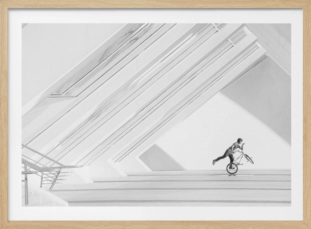 A minimalist black and white photograph in a silver frame, featuring a lone BMX rider performing a trick on the back wheel inside a stark, modern building with strong diagonal architectural lines. Poster