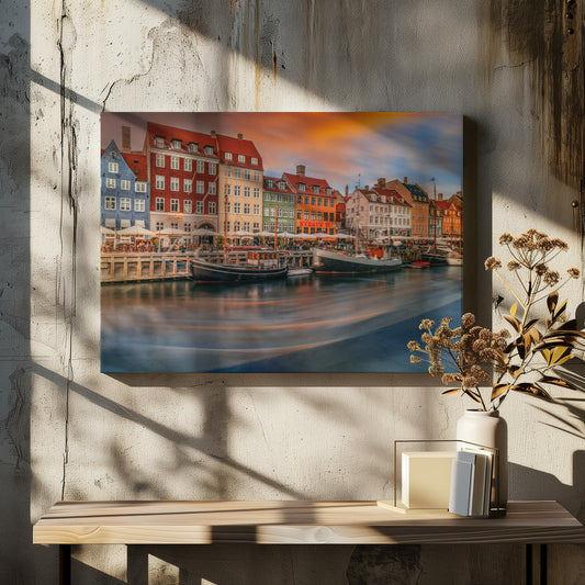 A long-exposure photograph of the colorful Nyhavn harbor in Copenhagen at sunset. Historic, brightly colored buildings line the canal, where two traditional wooden boats are moored. The sky is a dramatic blend of orange and blue, and the water is blurred, showing streaks of reflected light. Decor