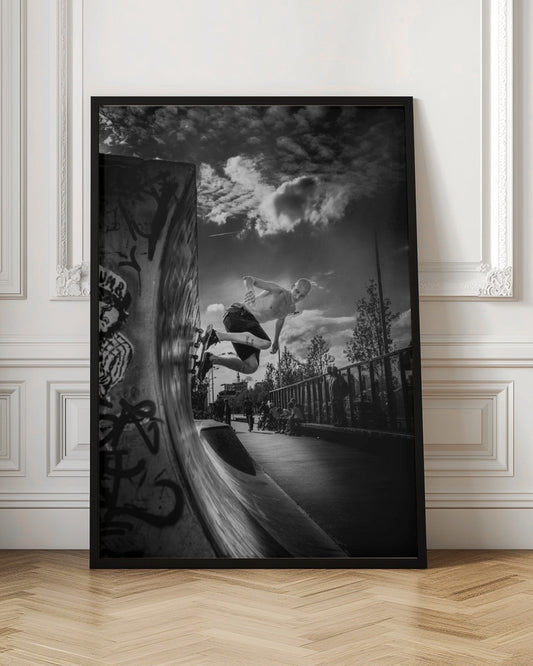 A dramatic black and white, low-angle action shot of a shirtless skateboarder suspended in mid-air at the peak of a graffiti-covered skate ramp, against a backdrop of a cloudy sky. Decor