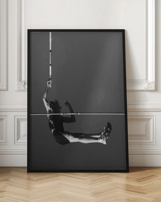 A dramatic black and white photograph of a male pole vaulter in mid-air, successfully clearing the horizontal bar. The athlete's body is parallel to the ground, with one arm still holding the vertical pole, against a solid gray sky. The image is enclosed in a simple black frame. Artwork