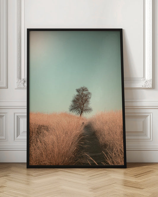A minimalist photograph of a single bare tree standing alone on a gentle slope in a field of golden, dried grass under a clear, pale teal sky. A narrow path winds its way through the tall grass toward the tree. Print