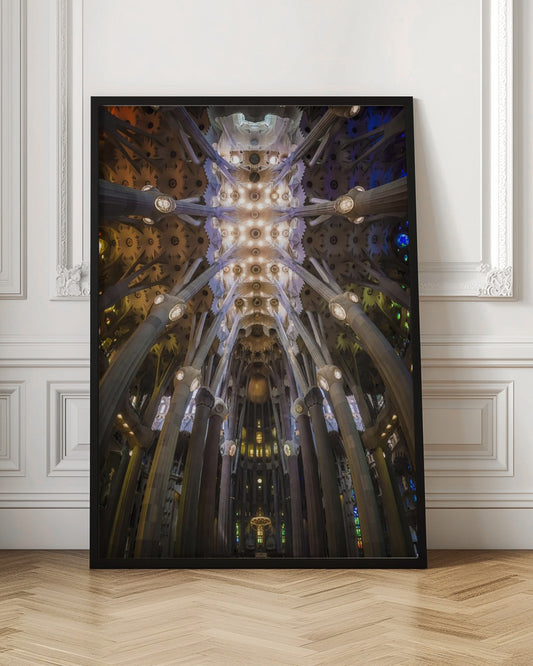 A low-angle, symmetrical photograph looking up at the incredibly ornate and complex vaulted ceiling of the Sagrada Familia Basilica. The massive stone columns branch out like trees, supporting the intricately detailed ceiling illuminated by warm, glowing lights and splashes of color from unseen stained glass windows. Poster