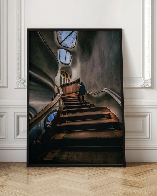 A low-angle view up a dark, winding wooden staircase inside a building with surreal, organic architecture. A person in a dark coat is walking up the stairs towards uniquely shaped windows that show a blue sky, contrasting with the dimly lit, textured gray walls. Print