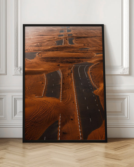 An aerial view of a multi-lane, black asphalt road being consumed by vast, rolling sand dunes of a deep orange color. The sand covers sections of the road, creating a surreal pattern of man-made lines and natural, windswept textures. Print