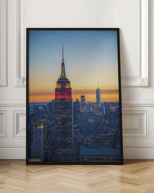 A vertical photograph of the New York City skyline at sunset, with the Empire State Building in the foreground. The building is illuminated with festive red and green lights on its upper floors, set against a sky that transitions from deep blue to a warm orange glow at the horizon. The surrounding cityscape, including the distant One World Trade Center, is bathed in the cool light of dusk, with scattered warm lights from building windows. Decor