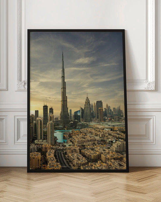 A framed photograph of the Dubai skyline at sunset, with the iconic Burj Khalifa skyscraper dominating the center. Surrounding it are numerous other modern high-rise buildings, a body of turquoise water, and a dense urban landscape in the foreground, all under a cloudy, golden-hued sky. Poster
