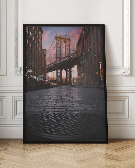 A low-angle photograph taken from a cobblestone street in Dumbo, Brooklyn, looking up towards the Manhattan Bridge at sunset. Red brick buildings frame the view on both sides of the street, with parked cars lining the left. A manhole cover is prominent in the foreground, and the sky is filled with warm pink and orange tones. Print