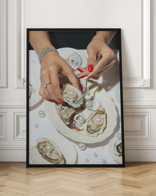 A close-up overhead shot of a woman's hands with vibrant red nails and elegant jewelry, using a fork to lift a large pearl from an oyster shell on a white plate. The plate and the surrounding white surface are adorned with numerous oysters filled with pearls of varying sizes, creating a luxurious and surreal scene. Print