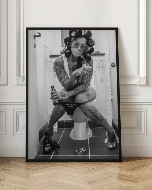 A black and white photo of a heavily tattooed woman with her hair in rollers sitting on a toilet. She wears large sunglasses, smokes a cigarette, holds a beer bottle, and has on black underwear that says 'CRAZY IN LOVE'. An ashtray and another beer bottle are on the floor. Artwork