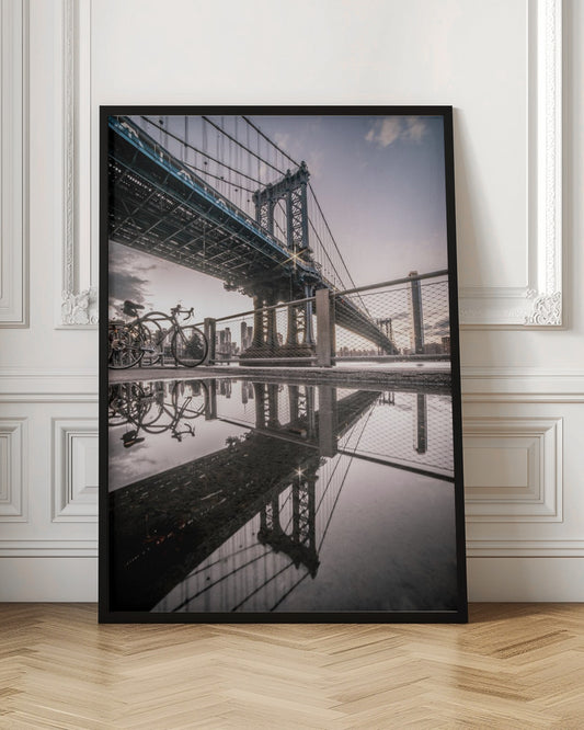 A dramatic low-angle photograph of the Manhattan Bridge, with its intricate steel structure and cables reflected in a large puddle on the pavement in the foreground. Two bicycles are parked near a fence on the left, with the New York City skyline visible in the distance under a softly lit, cloudy sky. Wall Art