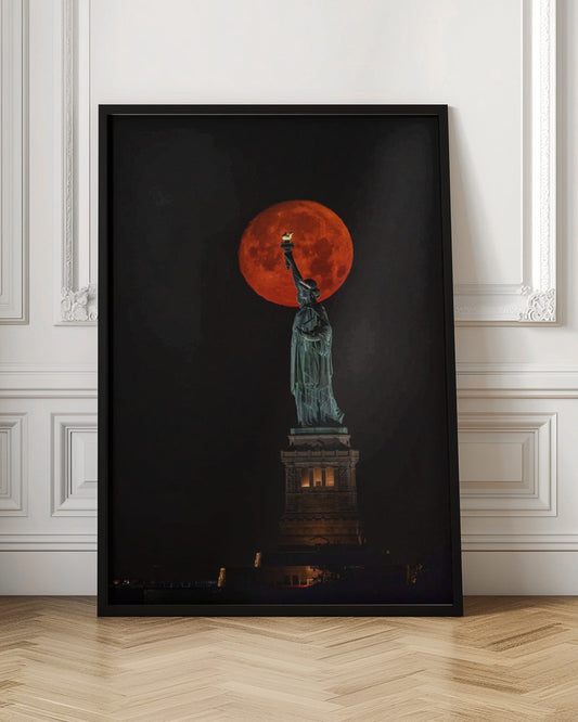 A striking night photograph of the Statue of Liberty perfectly silhouetted against a giant, vibrant red supermoon, which appears to be held aloft by the statue's torch against a deep black sky. Artwork