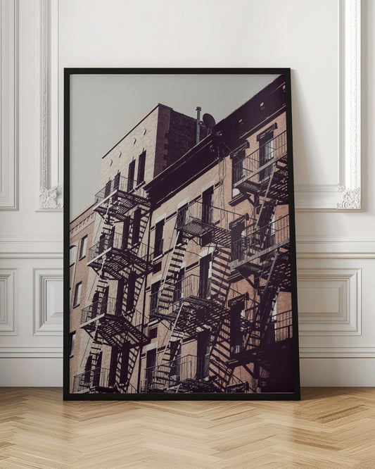 A low-angle photograph of a classic brick apartment building, with a complex pattern of black metal fire escapes casting long, dramatic shadows across the sunlit facade against a pale sky. Decor