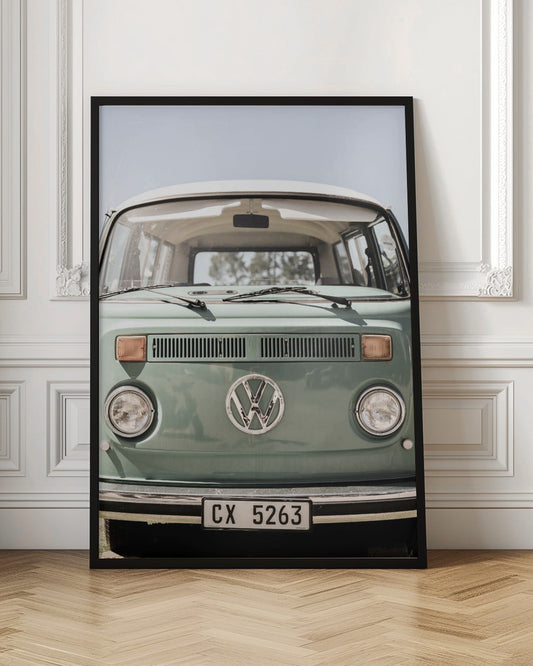 A close-up front view of a vintage sage green Volkswagen bus with a white roof. The iconic VW logo is on the front, with round headlights and a European license plate, set against a pale blue sky. Print