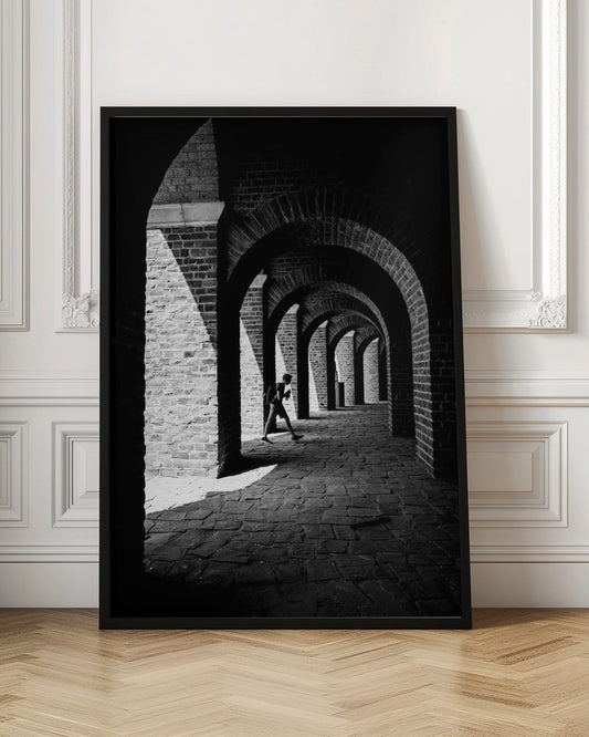 A dramatic black and white photograph of a person walking through a long brick arcade. Sunlight creates strong, contrasting shadows, highlighting the repeating arches that recede into the darkness, creating a powerful sense of depth and perspective. Decor