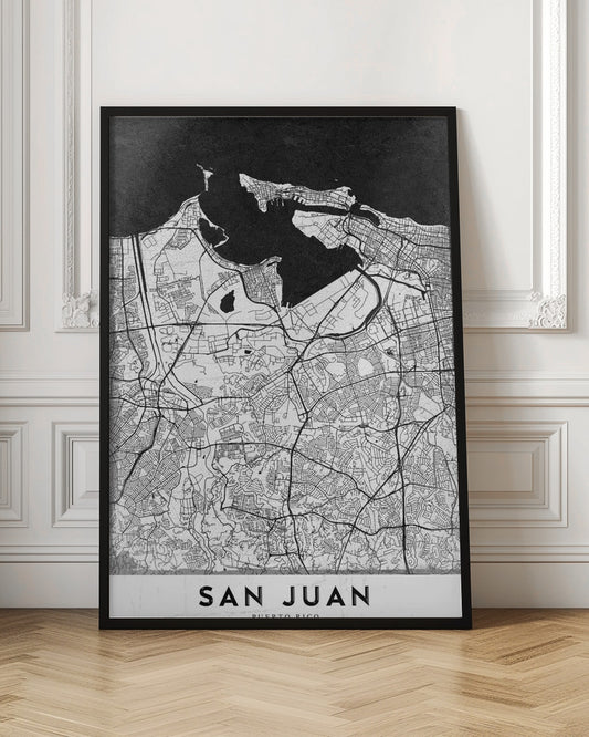 A minimalist black and white map of San Juan, Puerto Rico, shown as a framed poster. The land is depicted in white with intricate black lines for roads and streets, while the surrounding water is a solid, textured black. A white banner at the bottom features the text 'SAN JUAN' in a bold, black, sans-serif font. Artwork
