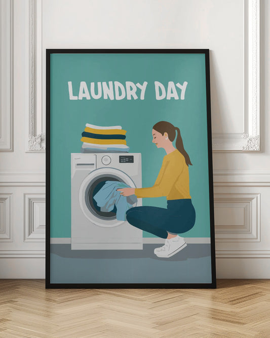 A framed illustration poster with the words 'LAUNDRY DAY' in white at the top. A woman with brown hair in a ponytail, wearing a yellow top and blue pants, crouches to load blue clothes into a white front-loading washing machine. A stack of folded laundry sits atop the machine against a teal background. Print