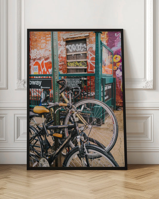 Several bicycles are locked to a green street pole in front of a red brick building covered in graffiti, with a city subway entrance visible in the background. Poster