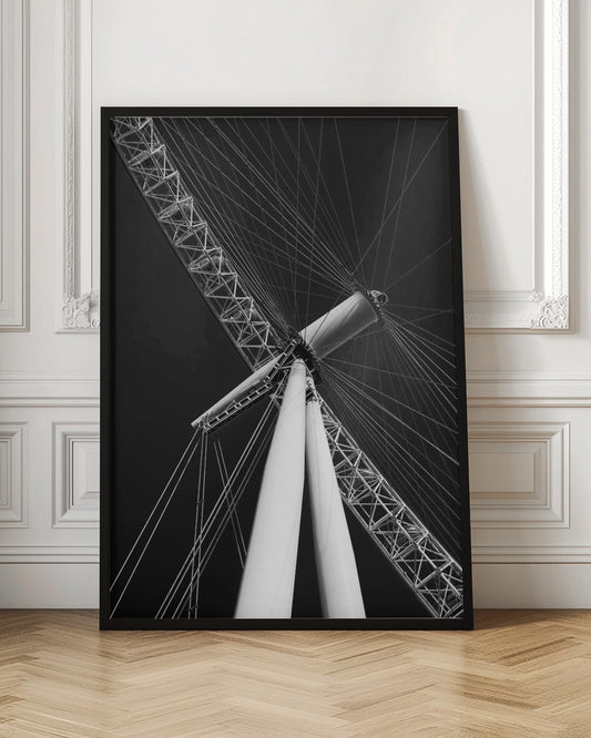 A dramatic low-angle, black and white photograph of a large Ferris wheel, focusing on the central hub and supporting structure. The image captures the white A-frame legs and the intricate lattice of the wheel's rim, with numerous tension cables radiating outwards against a solid black sky, creating a strong sense of perspective and geometric pattern. Artwork
