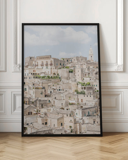 A vertical photograph of the ancient, stone city of Matera, Italy, with its dense collection of beige buildings and tiled roofs built up a hillside under a pale blue sky. A white bell tower stands out against the skyline. Poster