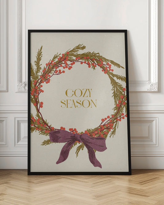 A vintage-style illustration of a festive wreath made of green boughs and red berries, tied with a large purple bow at the bottom. The text 'COZY SEASON' is written in gold inside the wreath on a cream-colored background, all enclosed in a thin black frame. Wall Art