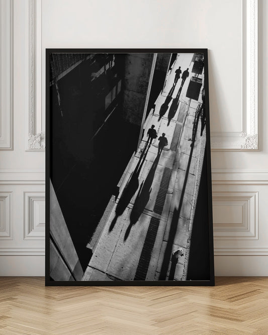 A dramatic high-angle, black and white photograph of several people walking on a sunlit sidewalk, casting long, stark shadows that stretch diagonally across the frame, contrasting with the deep black shadows of the adjacent building. Decor