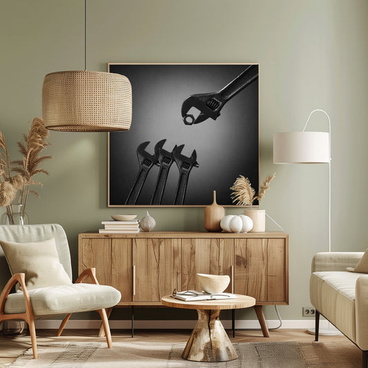 A conceptual black and white photograph in a light wood frame, depicting a large adjustable wrench holding a small nut above three smaller wrenches, which are angled upwards as if they were baby birds waiting to be fed. Print