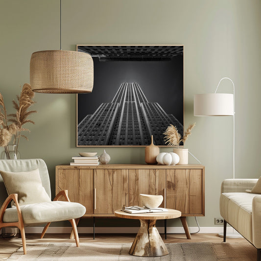 A dramatic, low-angle, black and white photograph of the Empire State Building, looking up from the street. The building's Art Deco lines stretch towards a dark grey sky, with another building visible at the very top of the frame, all enclosed in a light brown wood frame. Wall Art