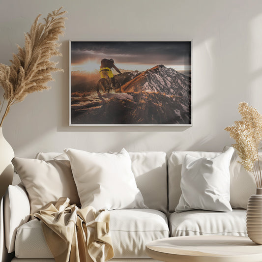 A mountain biker seen from behind rides along a narrow, snowy trail on a mountain ridge during a dramatic sunset, with the sun flaring brightly and a vast, snow-capped mountain range in the background. Decor