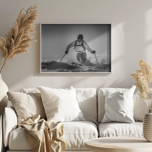 A black and white action photograph of a skier in full gear, leaning into a turn and spraying powder snow. The skier is backlit by the sun, creating a high-contrast, dramatic effect. Print