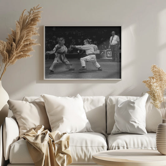 A black and white photograph of two young girls in a karate competition. They are wearing white gis, black belts, and boxing gloves. One girl is in the middle of throwing a punch at her opponent. In the background, a referee stands watching the match, and spectators are blurred in the stands. The image is presented within a silver-colored frame. Print