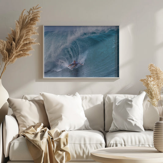 A dramatic action shot of a professional surfer skillfully navigating the inside of a massive, curling blue ocean wave, surrounded by white sea spray, all enclosed in a silver frame. Artwork
