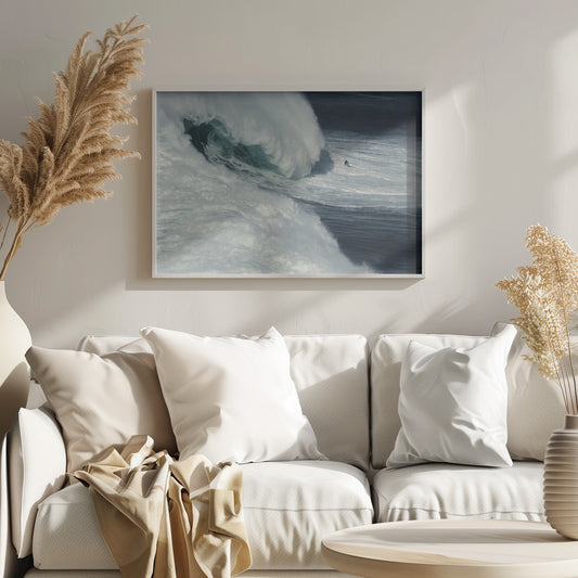 A framed aerial photograph captures a dramatic ocean scene where a lone surfer on a surfboard is dwarfed by an immense, powerful wave cresting nearby, conveying a sense of scale and adventure. Artwork