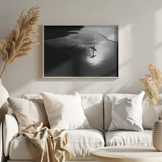 A high-contrast, black and white photograph of a silhouetted surfer carrying a surfboard and walking along the wet, reflective sand at the water's edge. Decor