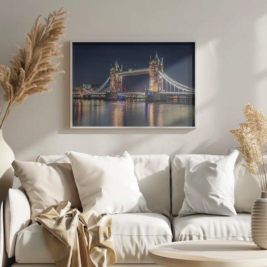 A stunning night photograph of London's iconic Tower Bridge, fully illuminated with golden and white lights that cast a shimmering reflection on the dark waters of the River Thames below, all set against a deep night sky and enclosed in a silver frame. Poster