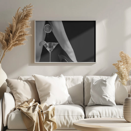 A framed, black and white photograph showing a woman's legs and a martini glass against a black background. A sliced citrus fruit is held between her thighs, with juice dripping towards the sugar-rimmed cocktail glass below. One foot is visible, wearing a black stiletto high heel. Artwork