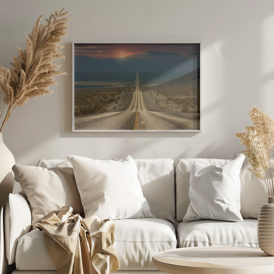 A high-angle view of a long, straight, and hilly road disappearing into the horizon of a vast desert valley at sunset, with mountains in the background under a warm, glowing sky, all enclosed in a silver frame. Poster