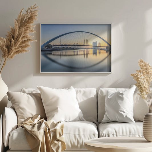 A framed photograph of the Dubai Water Canal Bridge at sunrise, with its large arch spanning across the water. The Dubai skyline, including the Burj Khalifa, is visible in the background, silhouetted against a golden and blue sky. The bridge and buildings create a perfect reflection on the calm surface of the water. Wall Art