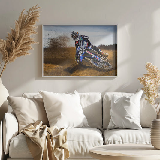 A motocross rider in full gear leans sharply into a turn on a dirt track, kicking up a large spray of mud and dirt from their blue and white Yamaha motorcycle. Artwork
