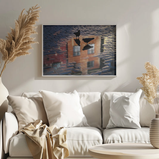 An artistic photograph of two silhouetted birds standing in a puddle on a wet cobblestone street. The puddle reflects an orange building with windows, and the birds' own reflections are visible beneath them, creating ripples in the water. The image is presented in a silver frame. Decor