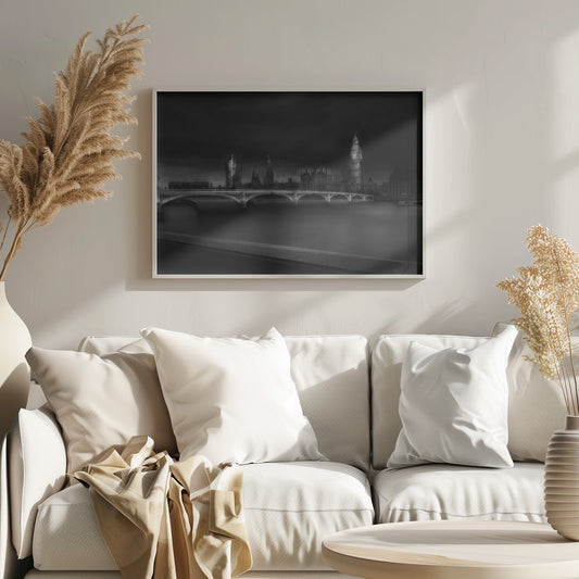 A moody, atmospheric black and white photograph of the Houses of Parliament and Big Ben in London, viewed from across the River Thames. The iconic buildings are softly illuminated against a dark, dramatic sky, with Westminster Bridge arching over the dark, smooth water in the foreground. The image is presented within a silver-colored frame. Artwork